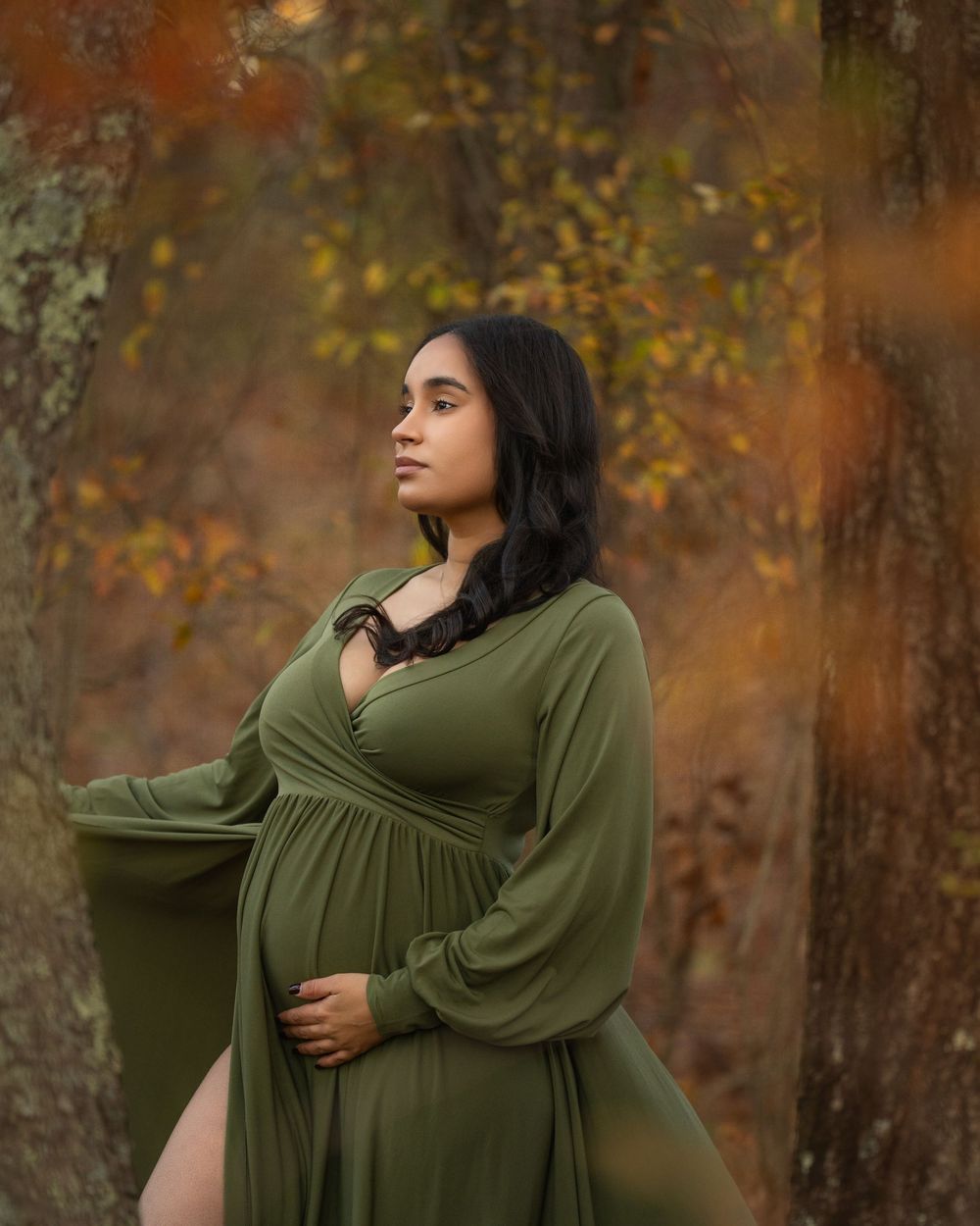 Pregnant woman in olive green maternity gown poses outdoors among autumn trees with golden foliage.