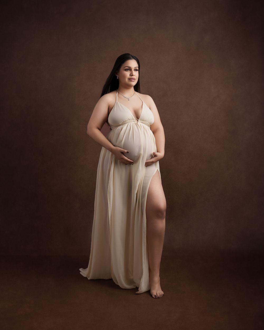 Maternity portrait in flowing cream dress against dark brown studio background.