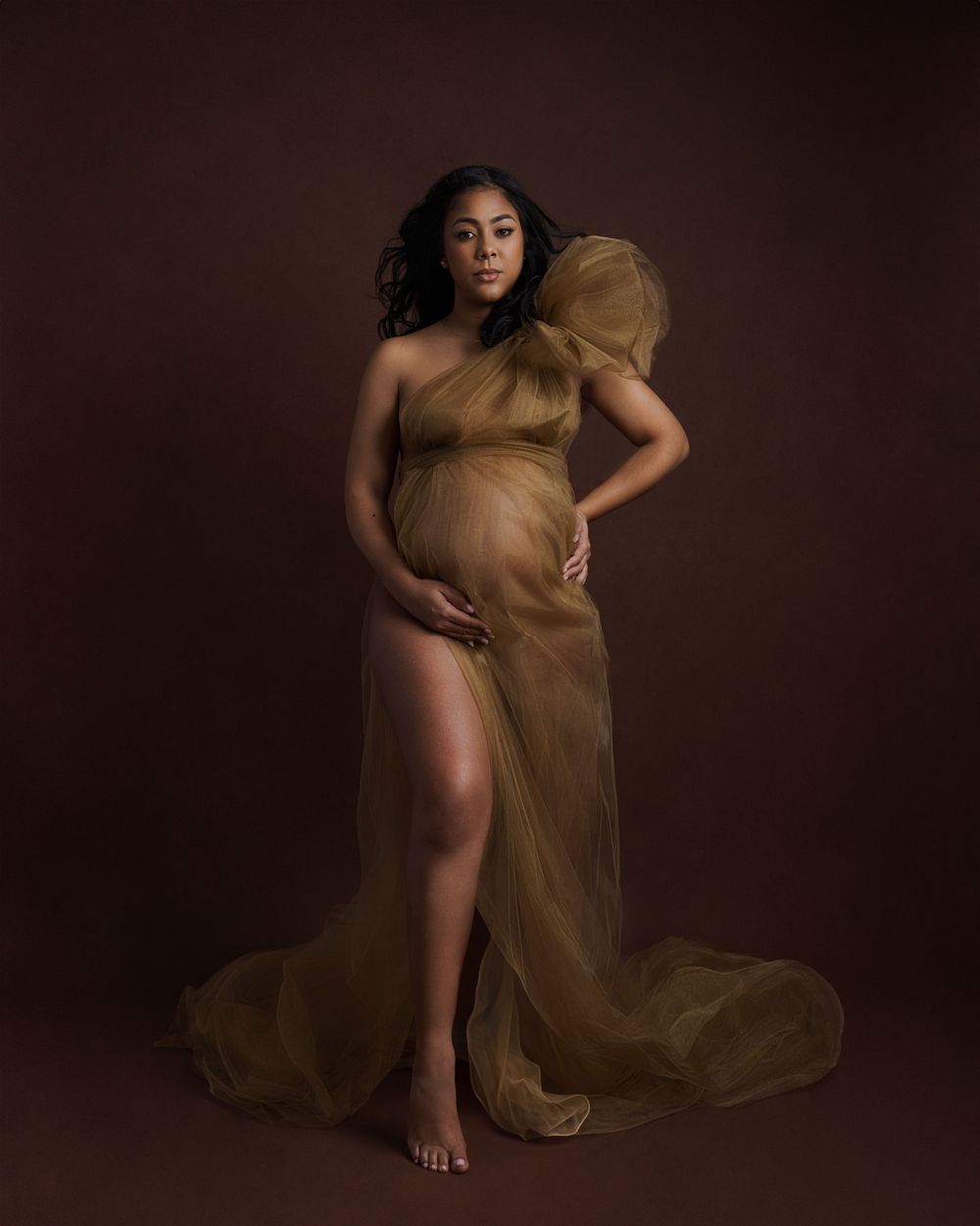 Dramatic maternity photoshoot featuring golden sheer fabric draping against dark backdrop.