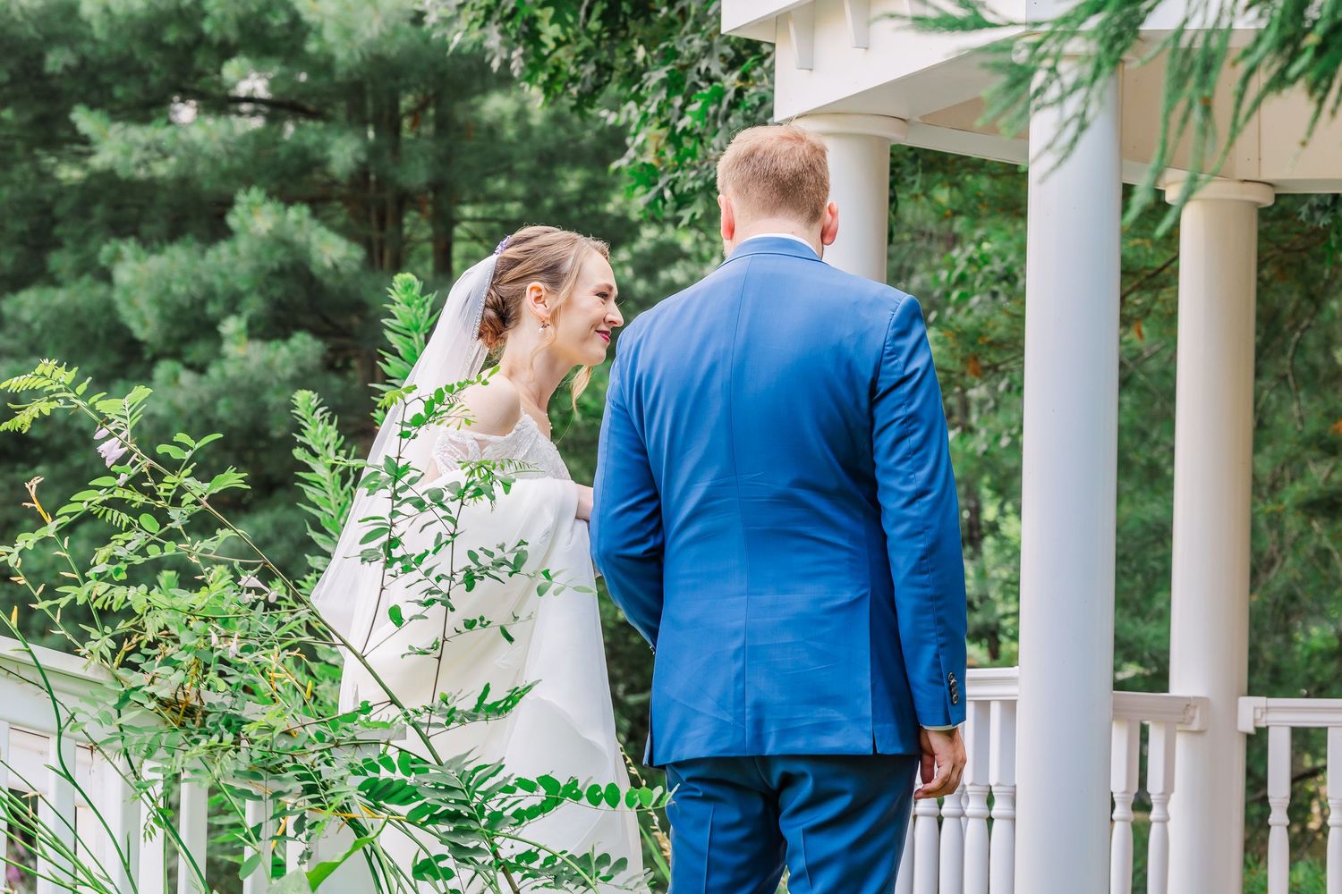 Elegant couple in formal attire stands on white porch near lush green foliage at outdoor wedding venue.