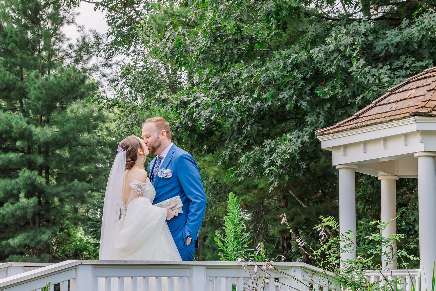 A couple shares a romantic moment on a white deck railing with a garden gazebo and lush trees in the background.