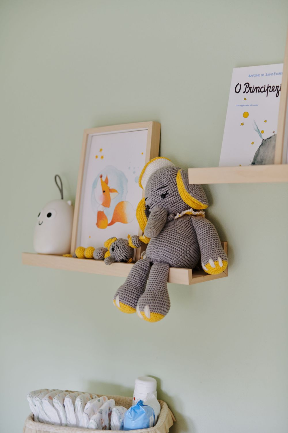 A nursery shelf decorated with a crocheted elephant toy, fox print, and Little Prince book on a green wall.