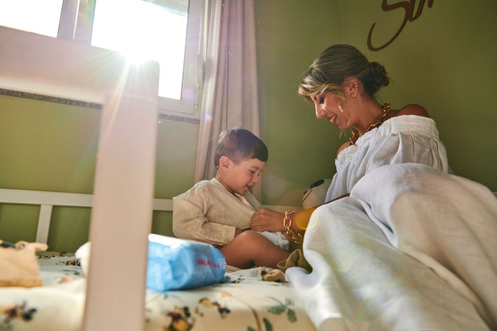 A mother sits on a bed with her young toddler in a green nursery room by a bright window.