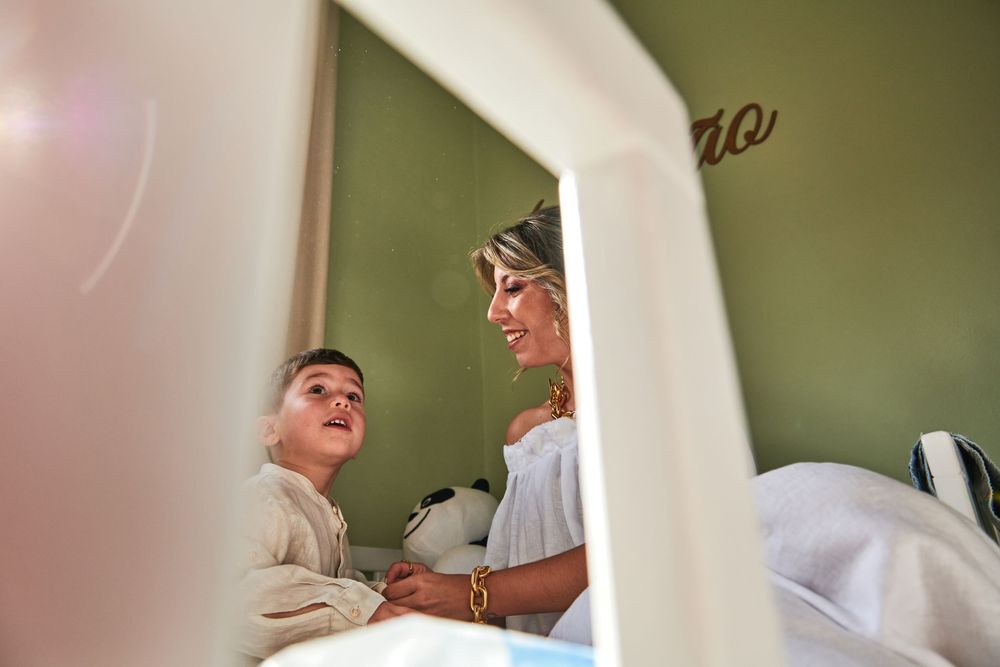 A mother smiles warmly at her young child in a green nursery room during an intimate family moment.