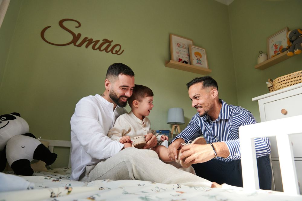 Two fathers sit on a bed with their toddler in a green nursery named Simao, sharing a joyful moment.
