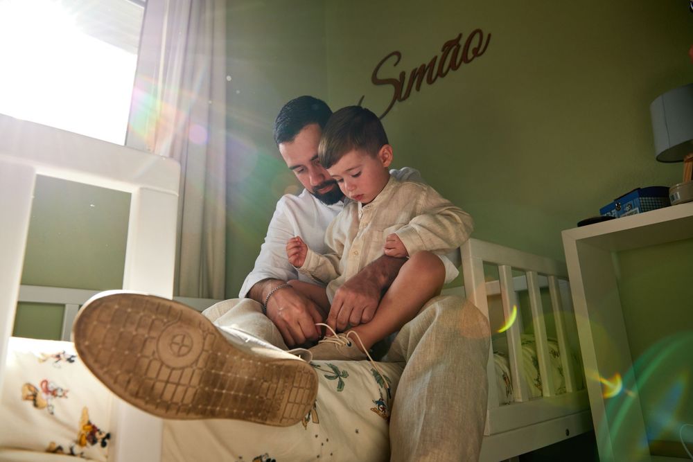 A father helps his young son put on shoes while sitting together on a bed in a green nursery room.