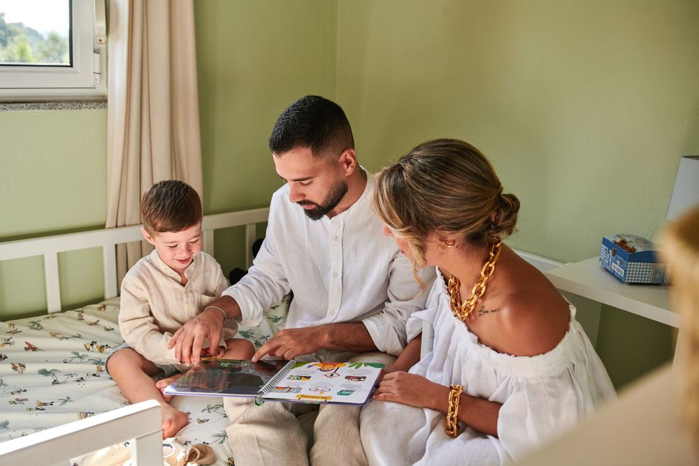 A family of three reads a picture book together on a bed in a cozy green childrens bedroom.