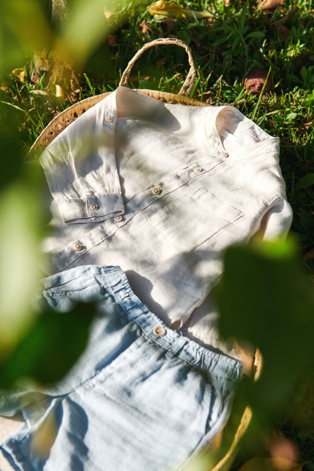 A white linen shirt and light blue pants laid on a wicker tray in a sunny garden setting.