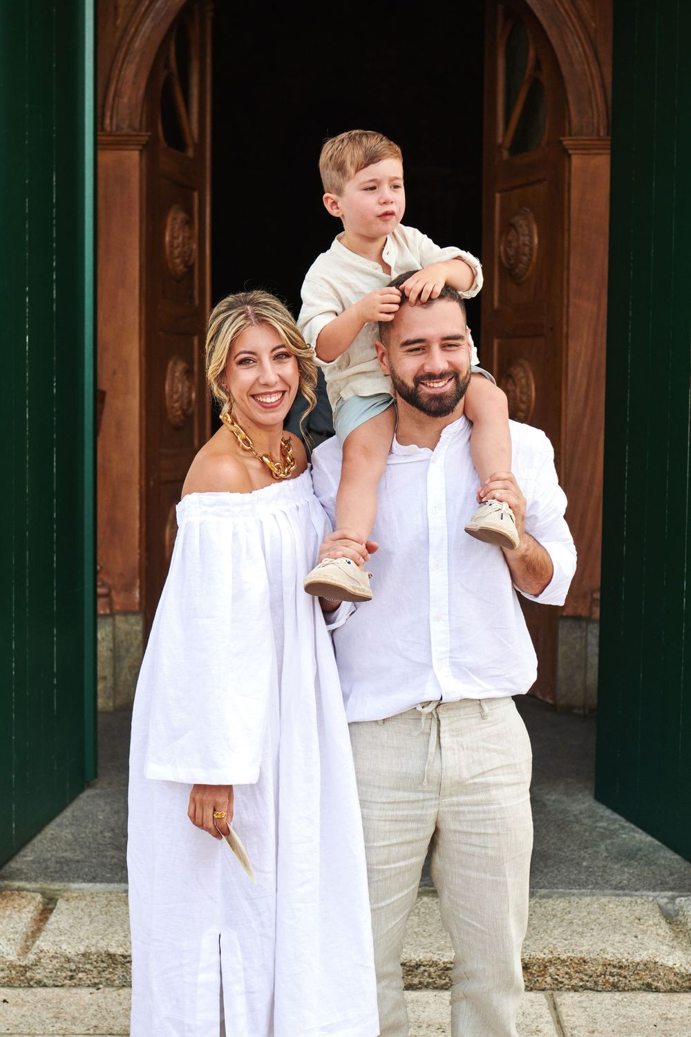 A smiling family in white outfits poses outside ornate wooden church doors with child on fathers shoulders.