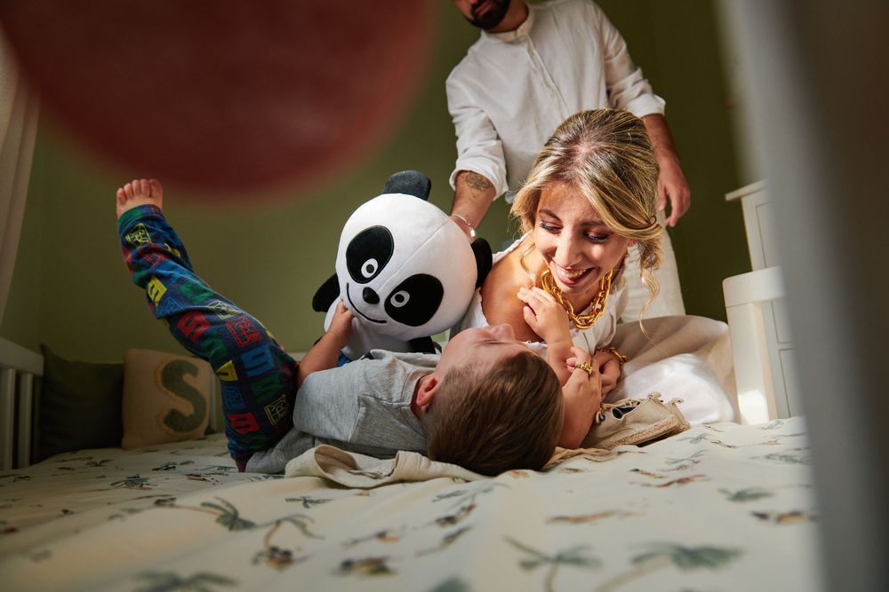 A mother plays and laughs with her toddler on a bed while an older child holds a panda stuffed toy.