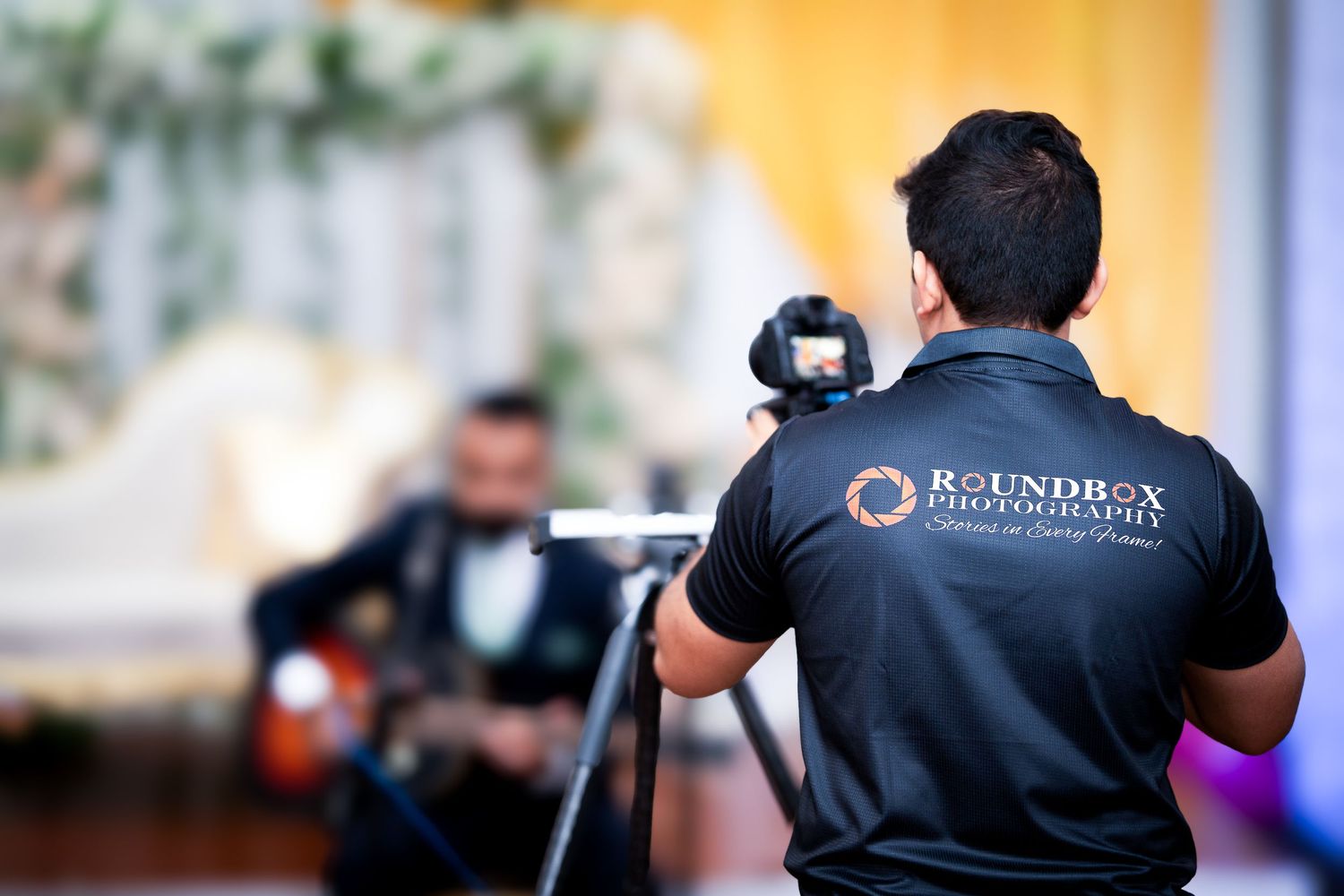 Videographer wearing Roundbox photography shirt films event with professional camera at indoor venue.