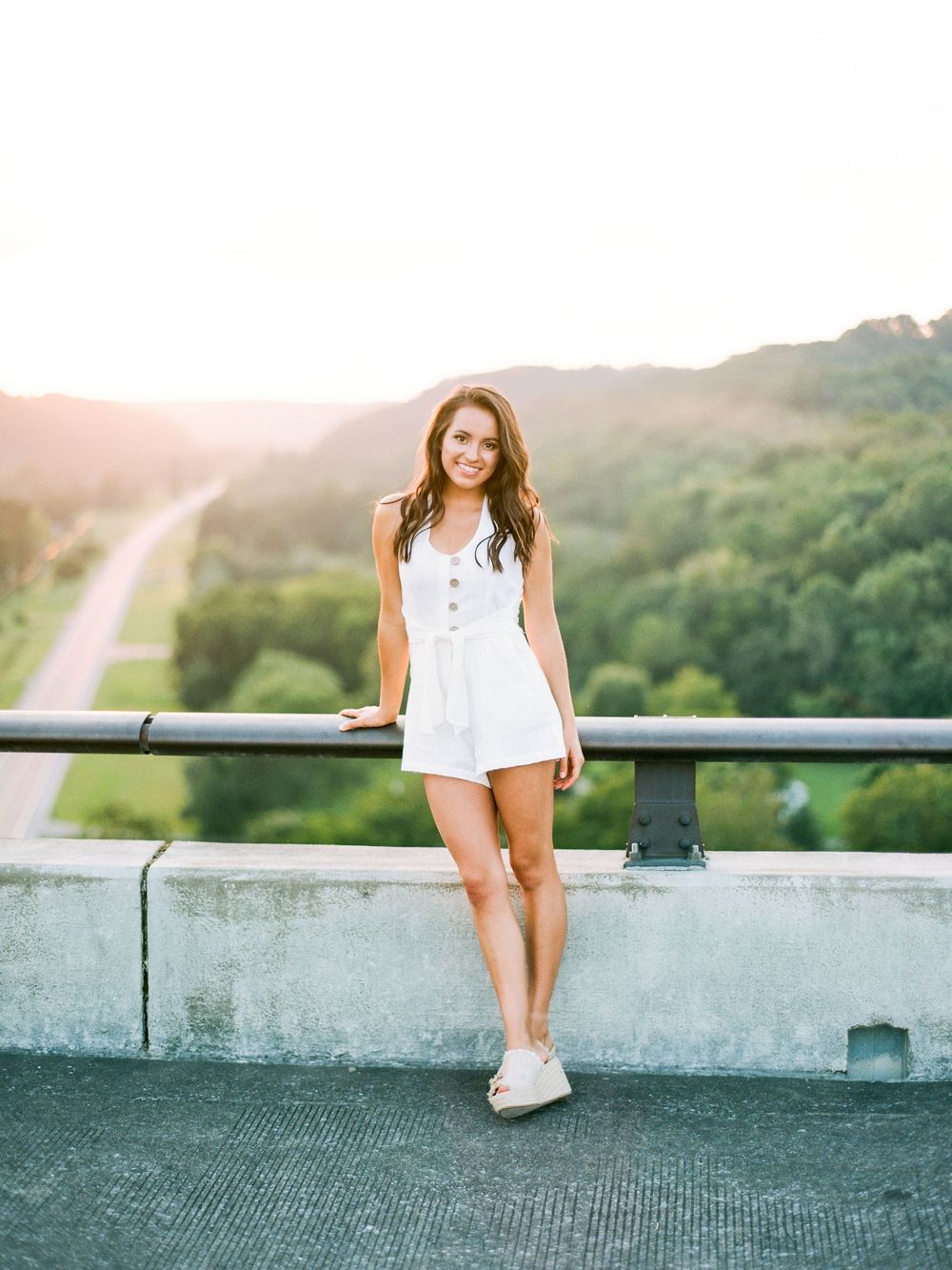 Person in a white summer romper leans against railing on scenic overlook at sunset with winding road in background.