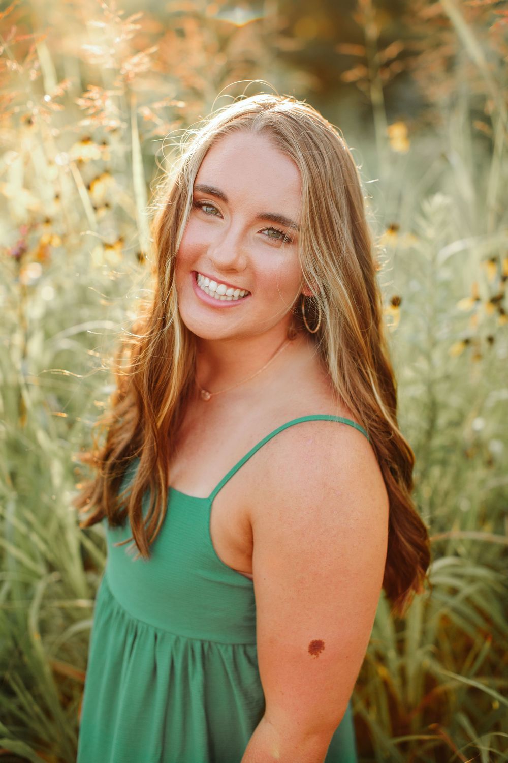 Portrait of someone wearing a green sundress smiling while posing in tall grass during golden hour lighting.