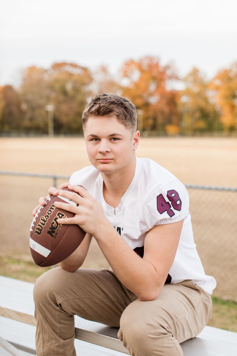 Young athlete in white jersey number 49 sits on bleachers holding football during autumn season.