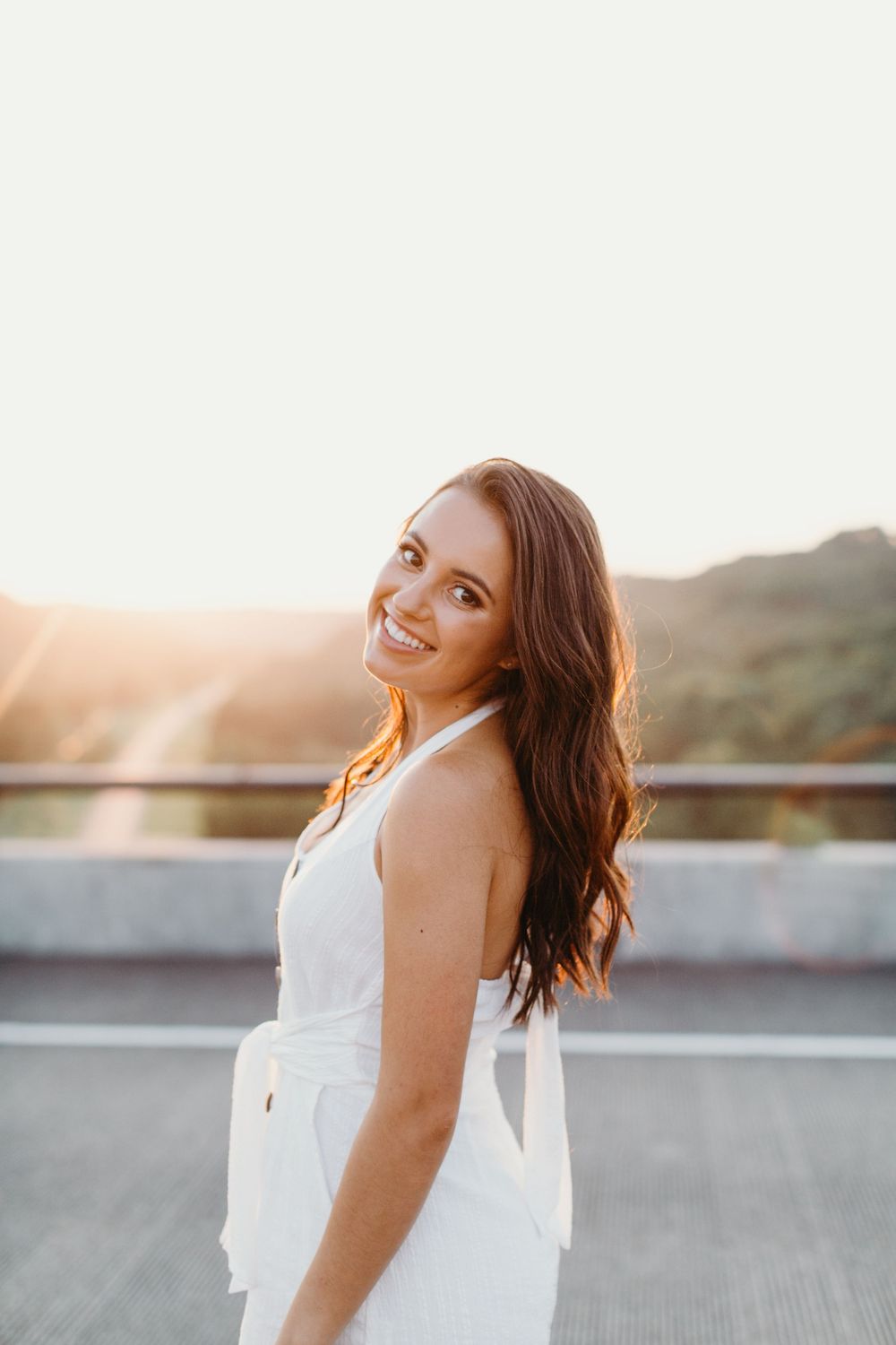 A person in a white dress smiles joyfully during sunset on a rooftop with mountains in the background.
