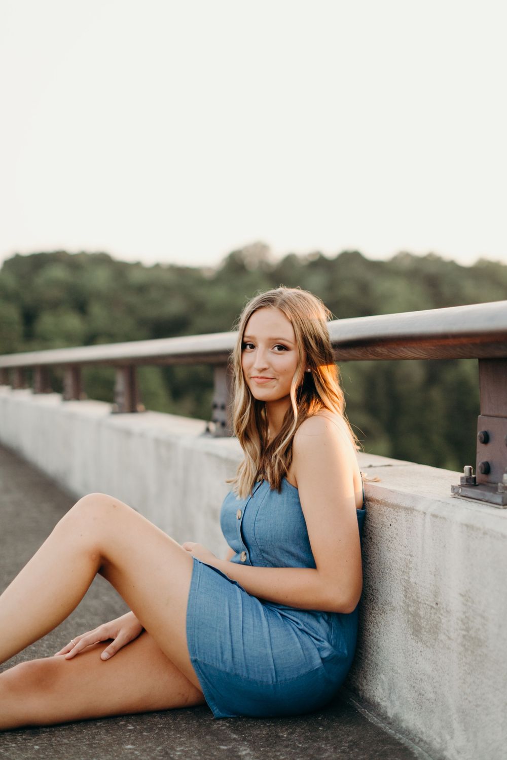 Young woman in blue dress sitting on concrete ledge against railing during sunset photoshoot.