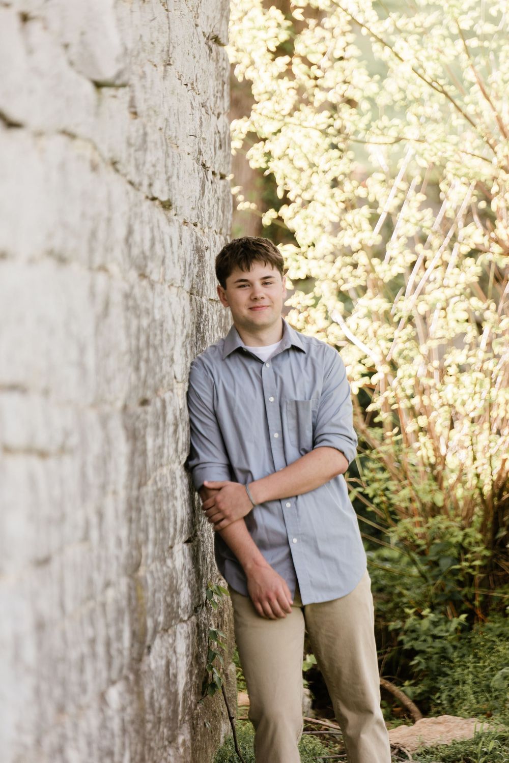 Person in grey button-down shirt and khaki pants poses against weathered wall with blooming foliage in background.
