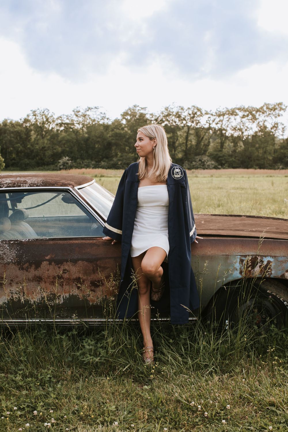 A rustic fashion photo shoot features a white dress and long coat posed against a vintage rusty car in a grassy field.