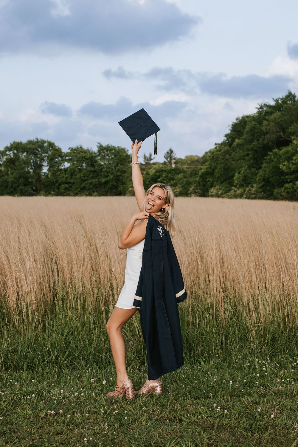 Graduate celebrates in a field wearing graduation gown while holding cap up in the air on sunny day with wildflowers.