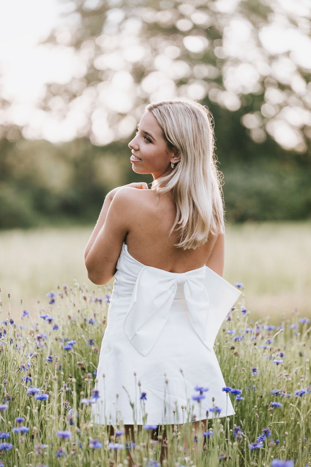 A woman in a white strapless dress with bow detail stands in a field of blooming purple cornflowers at sunset.