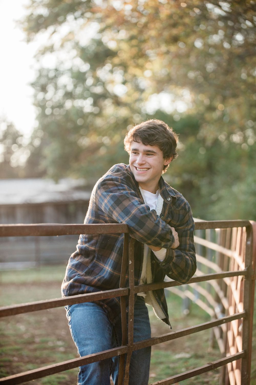 Person wearing a plaid flannel shirt and jeans leans on rustic fence post during golden hour in outdoor rural setting.