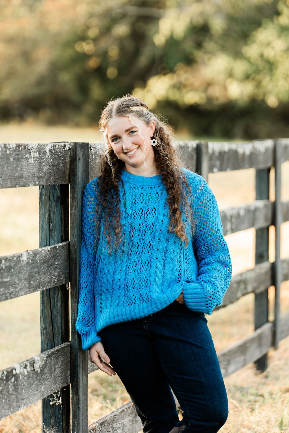 A maternity photo shoot at sunset shows someone in a bright blue sweater standing by a rustic wooden fence.