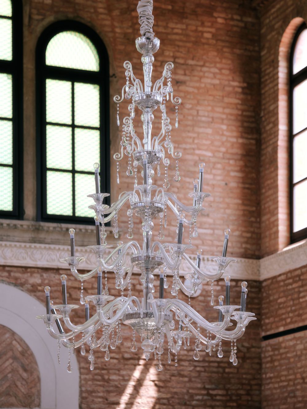 Stunning crystal chandelier hanging in a sunlit brick room with arched windows. Photoshoot by Venice Photographer Alina Indi