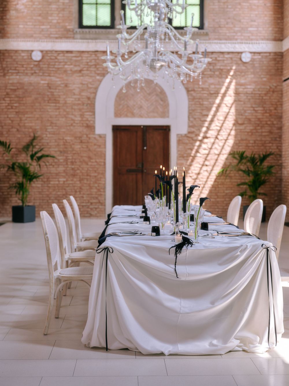 Elegant wedding reception setup with crystal chandelier and white draped table in brick venue. Photoshoot by Venice Photographer Alina Indi
