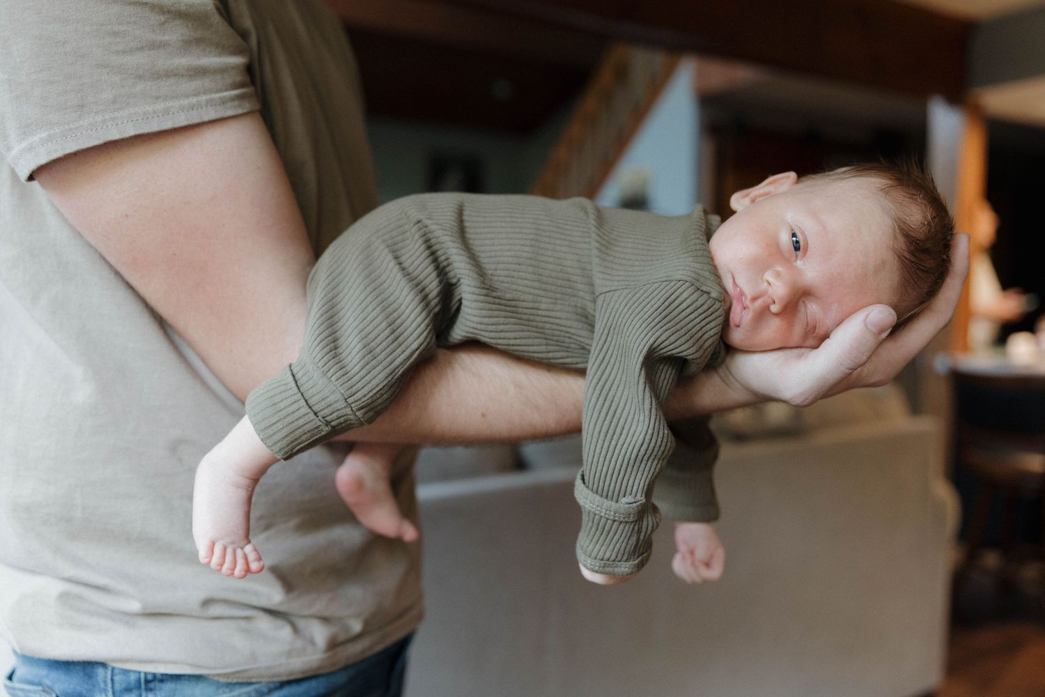 A sleeping baby wearing green pajamas rests peacefully on someone's arm in a soft, warm indoor setting.