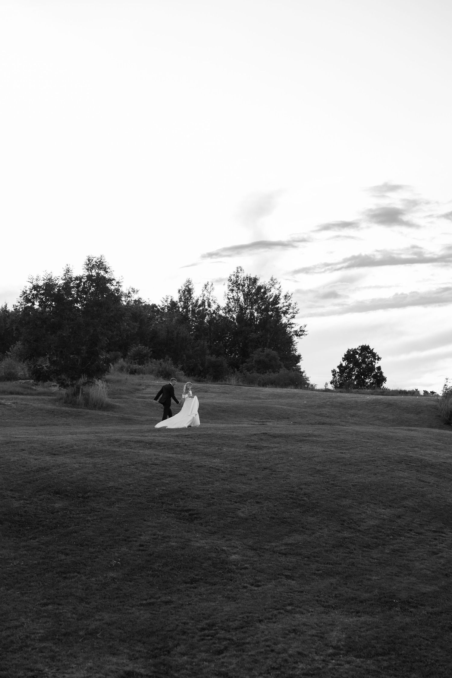 Two silhouettes stand atop a green hillside with trees at sunset creating a romantic panoramic scene.
