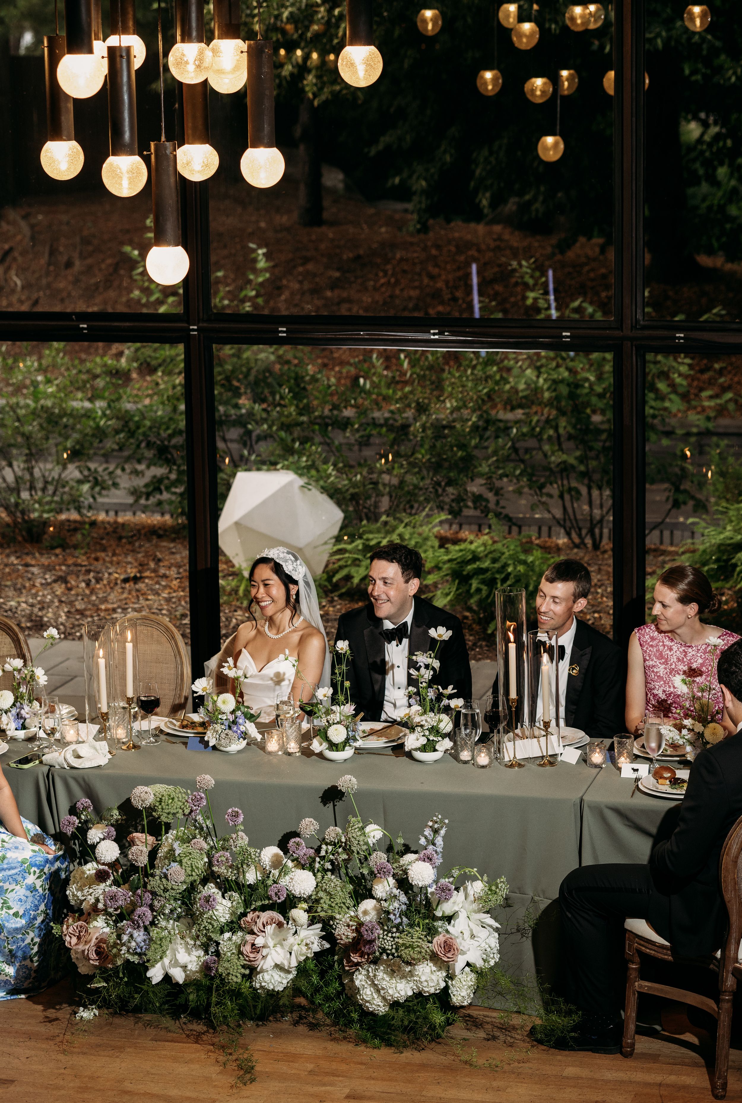 Wedding reception table decorated with lush floral arrangements and lanterns hanging overhead in an elegant rustic setting.