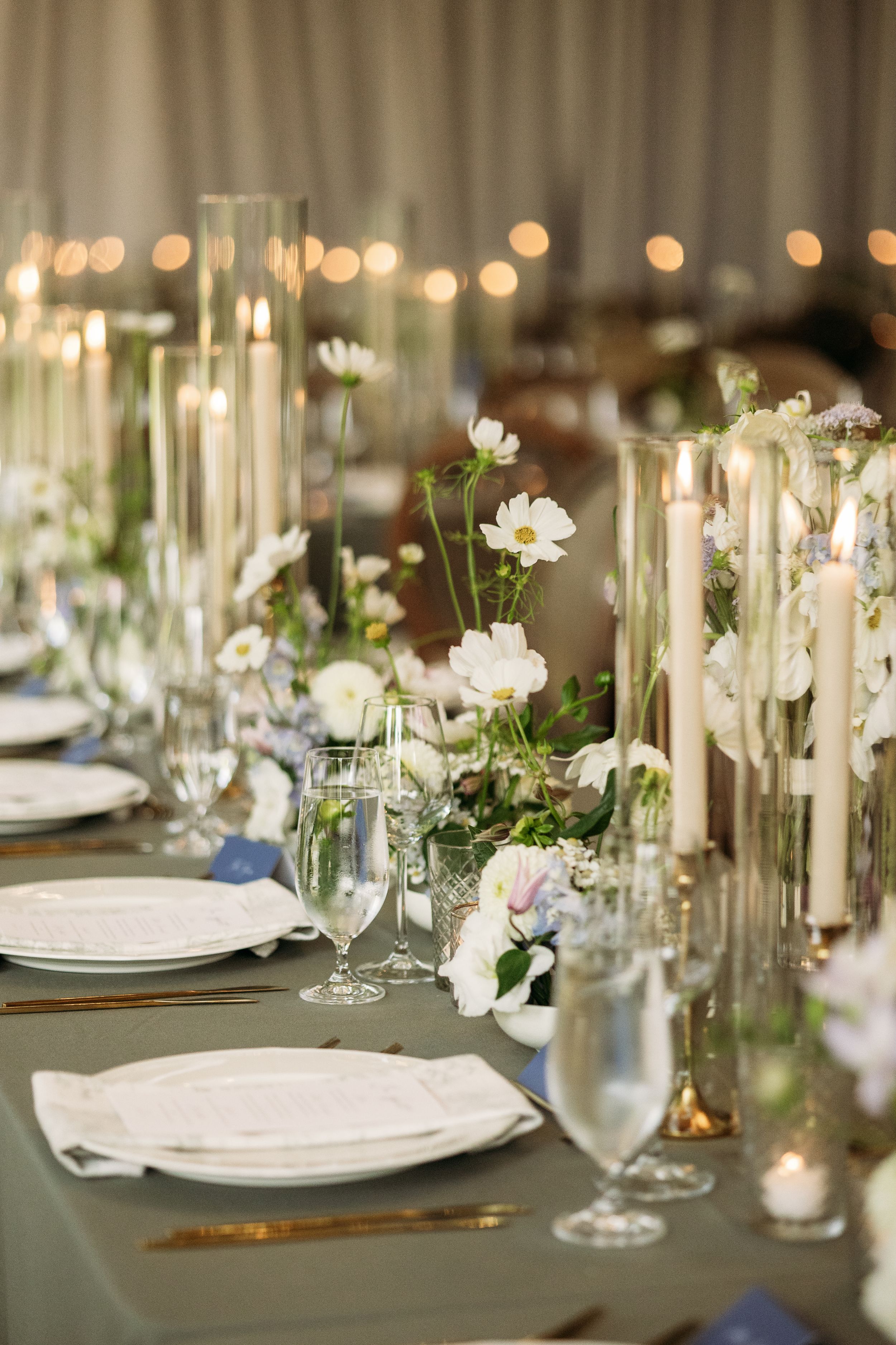 Elegant wedding table setting with tall glass candle holders, white flowers and sage green linens under twinkling lights.