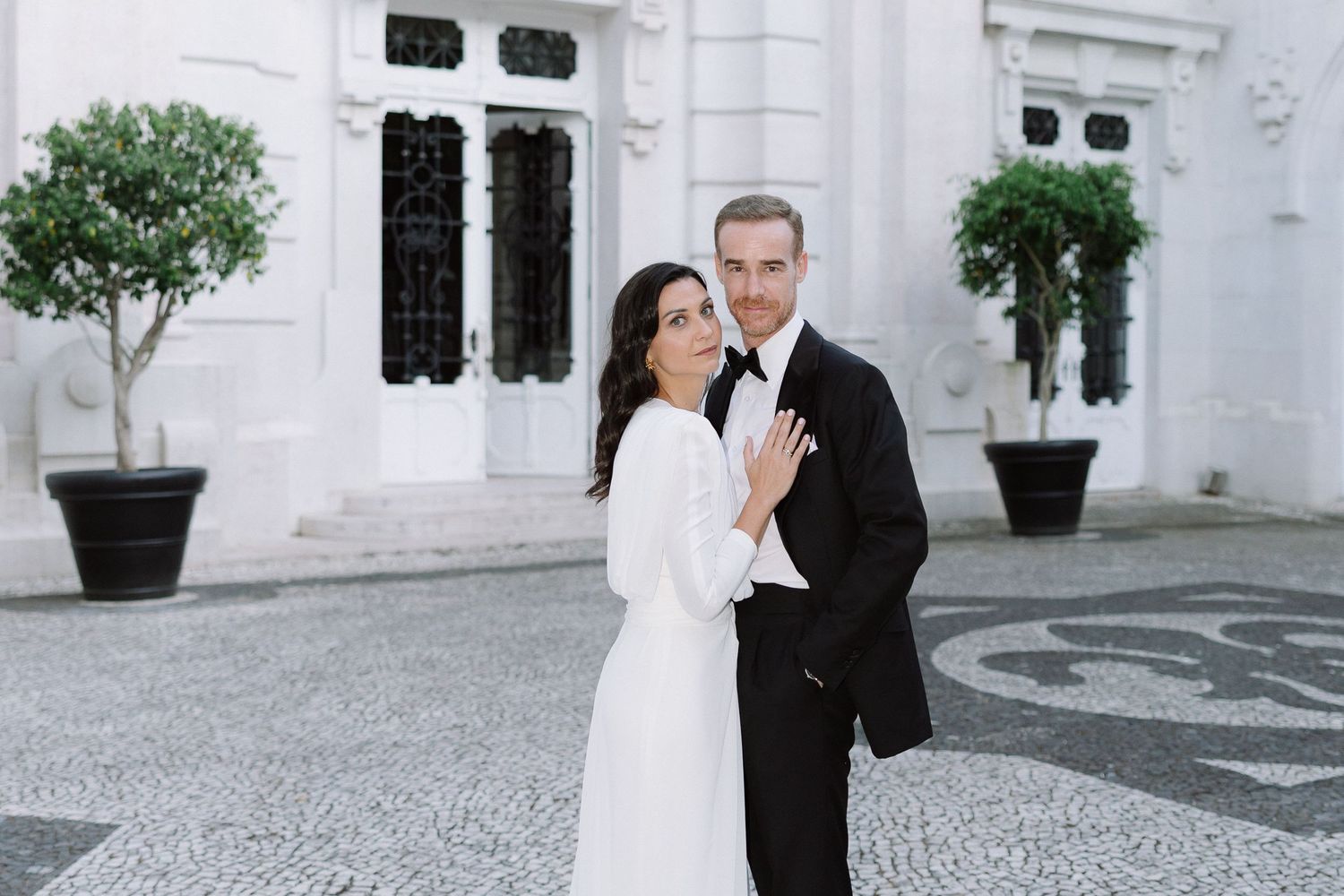 Elegant couple in formal attire walking down ornate hallway with light grey walls and gold framed artwork.
