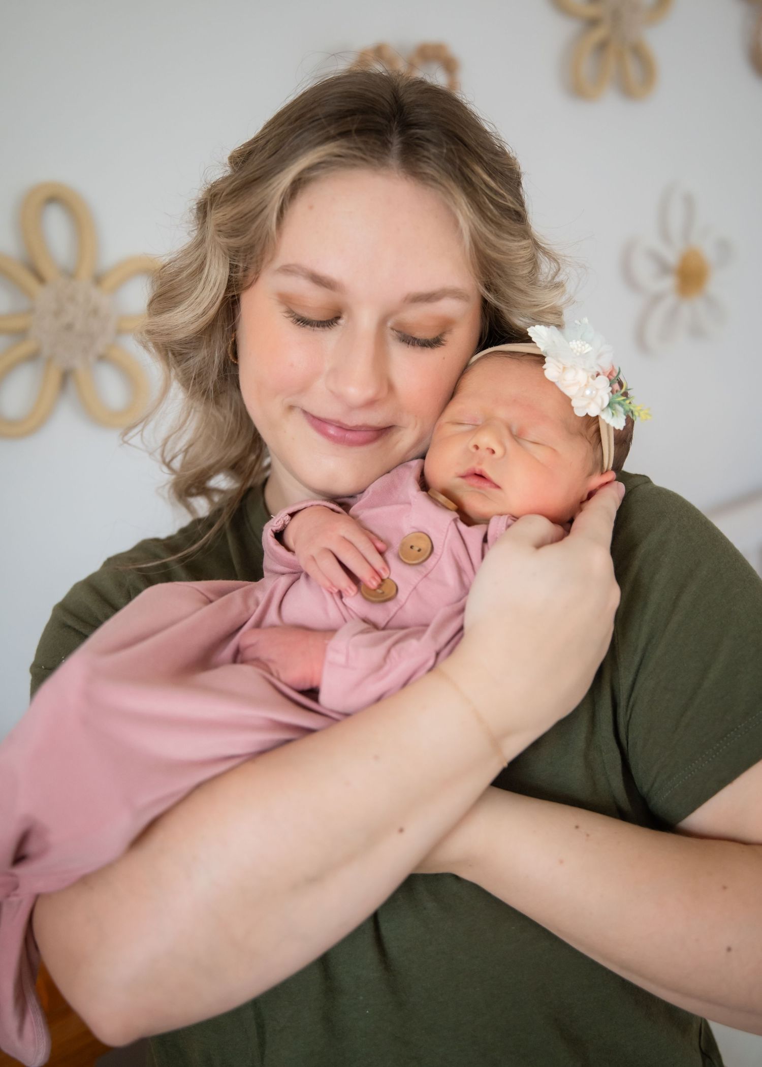 A woman in olive green shirt and jeans cradles and gazes lovingly at her newborn baby in front of a white crib.