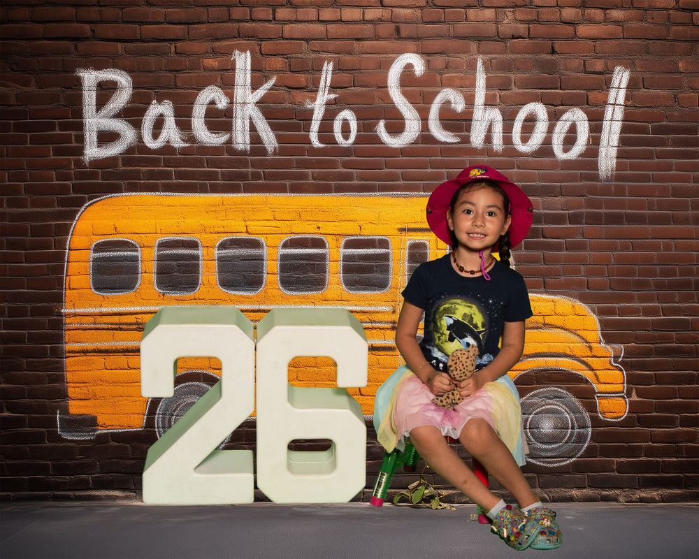 A child sits in front of a brick wall with painted yellow school bus and 'Back to School' text for a first day photo.