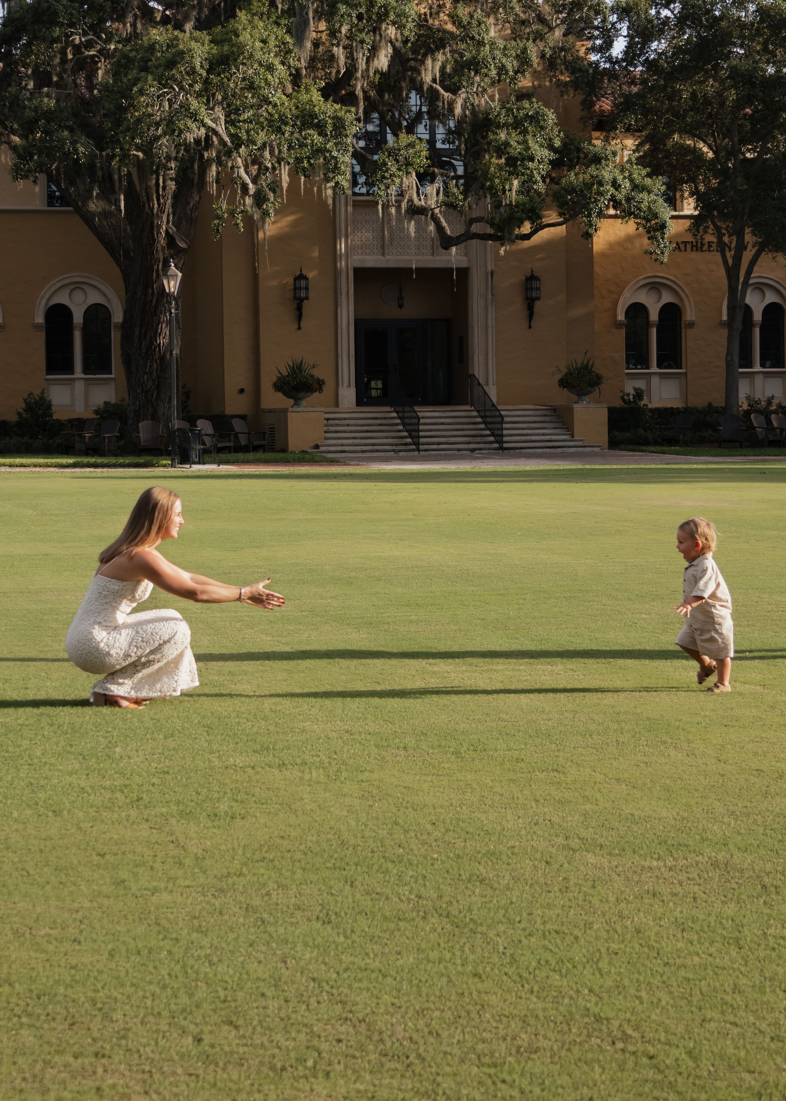 People in white dresses play and interact on a large green lawn near a building.