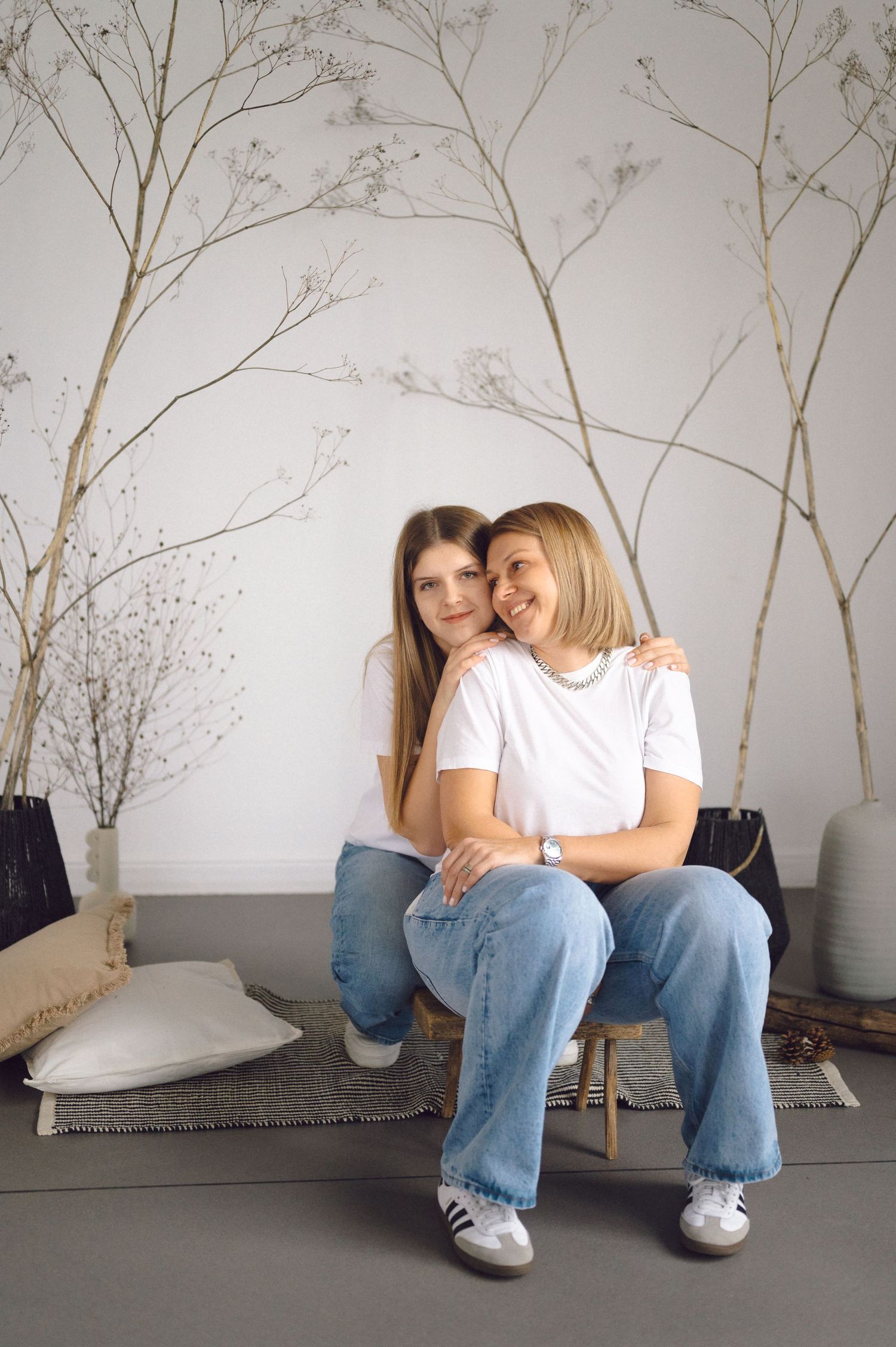 Two people in matching white shirts and jeans sit close together on a bench against a wall with bare branches.