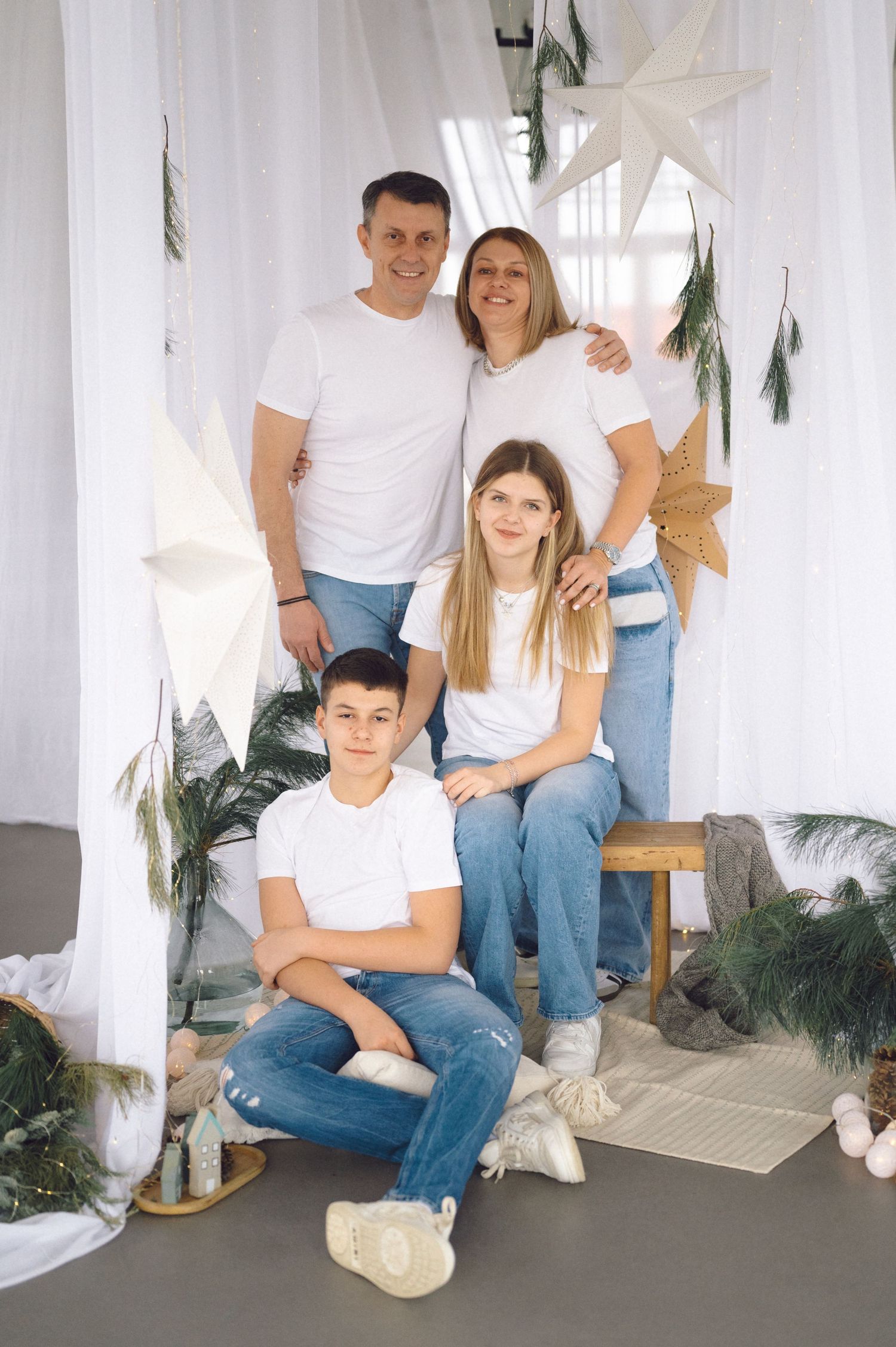 A family sits together in matching white shirts and jeans against a light decorated background with greenery accents.
