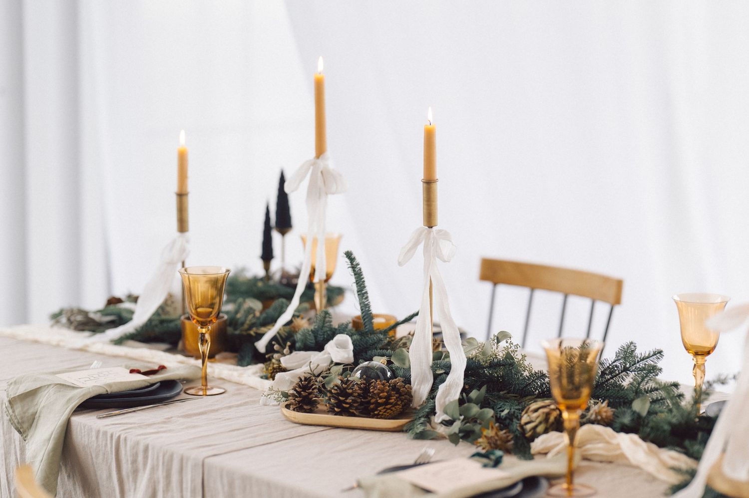 Elegant holiday table setting with white candles, amber glasses, and evergreen garland centerpiece on linen tablecloth.