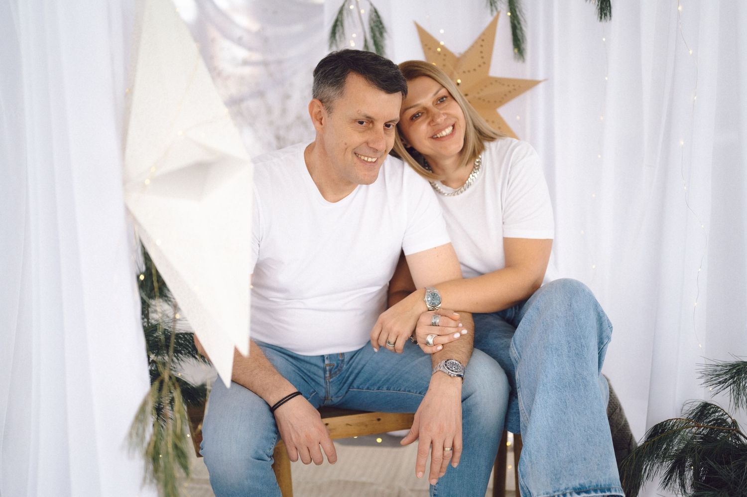 Couple in white t-shirts and blue jeans sitting together on wooden box with Christmas decor and star ornament.