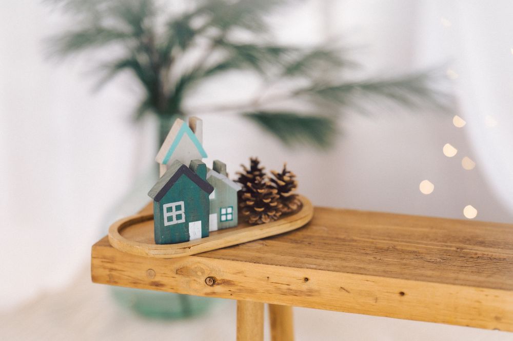 A small teal house decoration sits on a wooden shelf with pinecones and blurred bokeh lights in the background.
