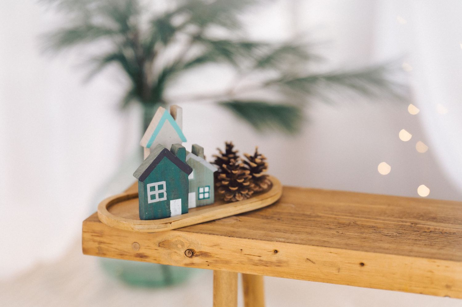A small teal house decoration sits on a wooden shelf with pinecones and blurred bokeh lights in the background.