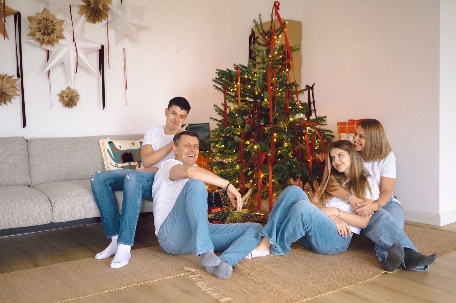 A group relaxes in a living room with a decorated Christmas tree, surrounded by holiday decor and wall ornaments.