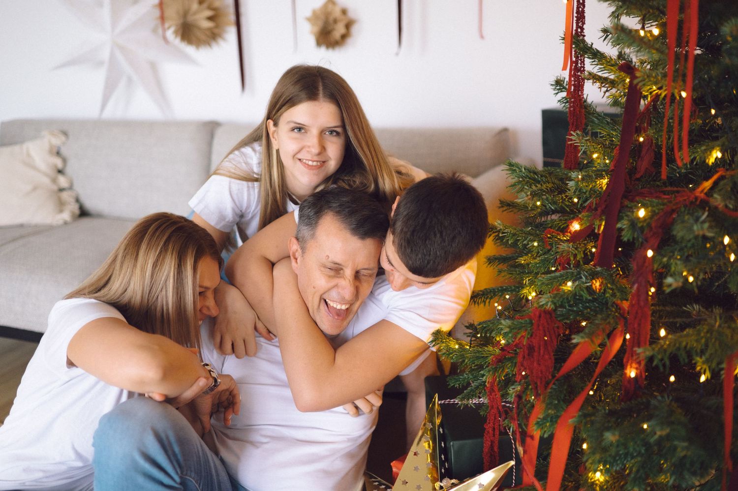 Family cuddles together on a couch beside a decorated Christmas tree with twinkling lights and red ornaments.