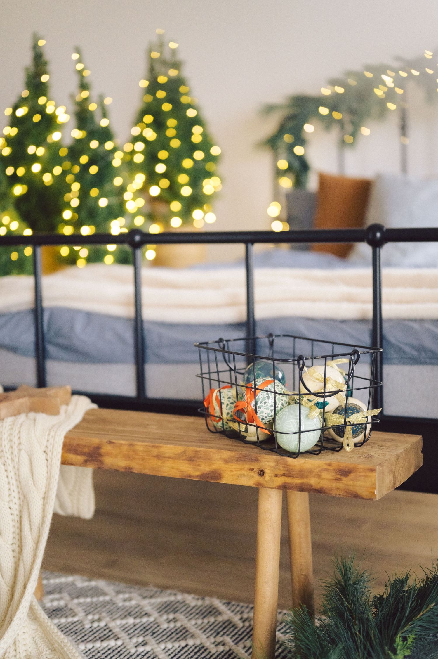 Wire basket filled with ornaments sits on a wooden bench with cozy holiday decor and lit Christmas trees in background.