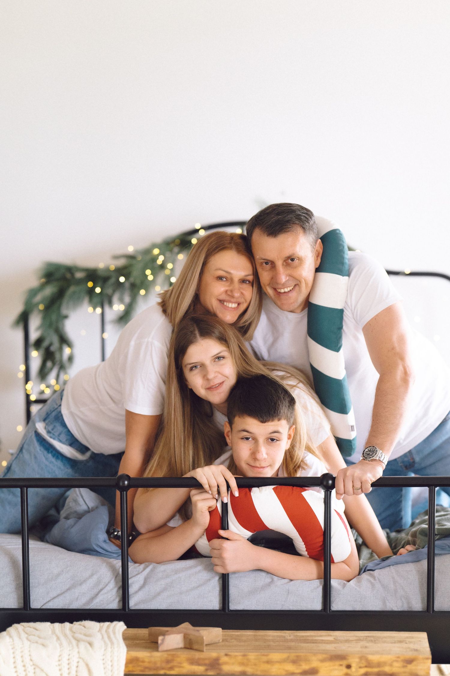 Happy family wearing white shirts lying together on a black metal bed frame, posing playfully for the camera.