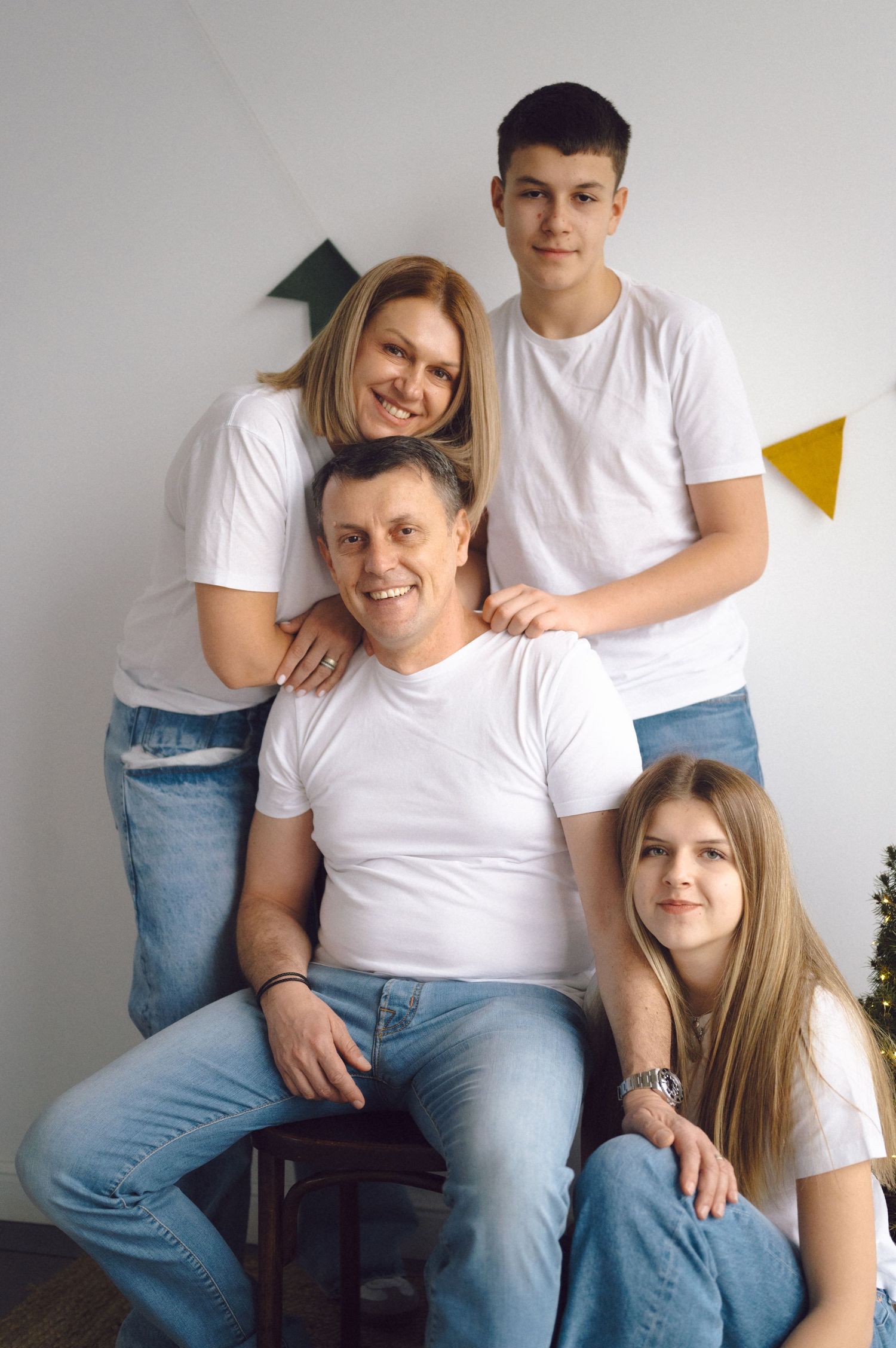 Family of four wearing matching white t-shirts and blue jeans pose together against a gray wall with colorful triangles.