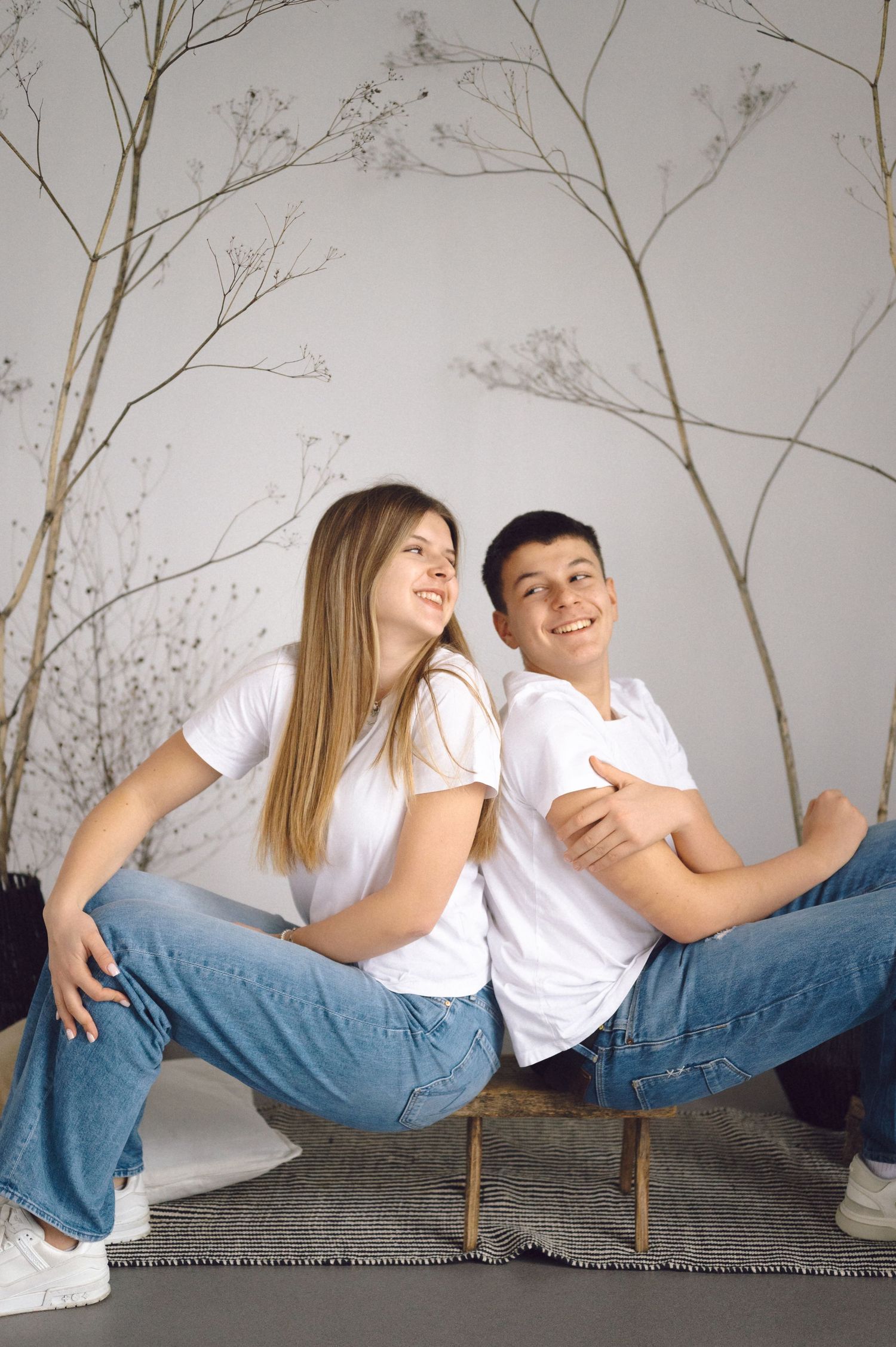 A couple in matching white shirts and blue jeans sits back to back, laughing together against a gray wall with branch decor.