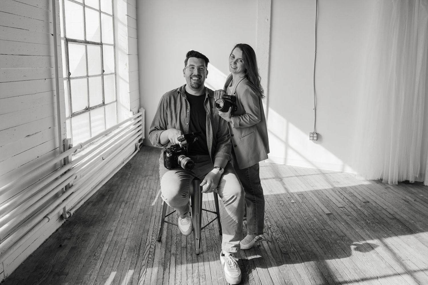 A couple laughs together while posing with a camera in a bright sunlit room with wooden floors.