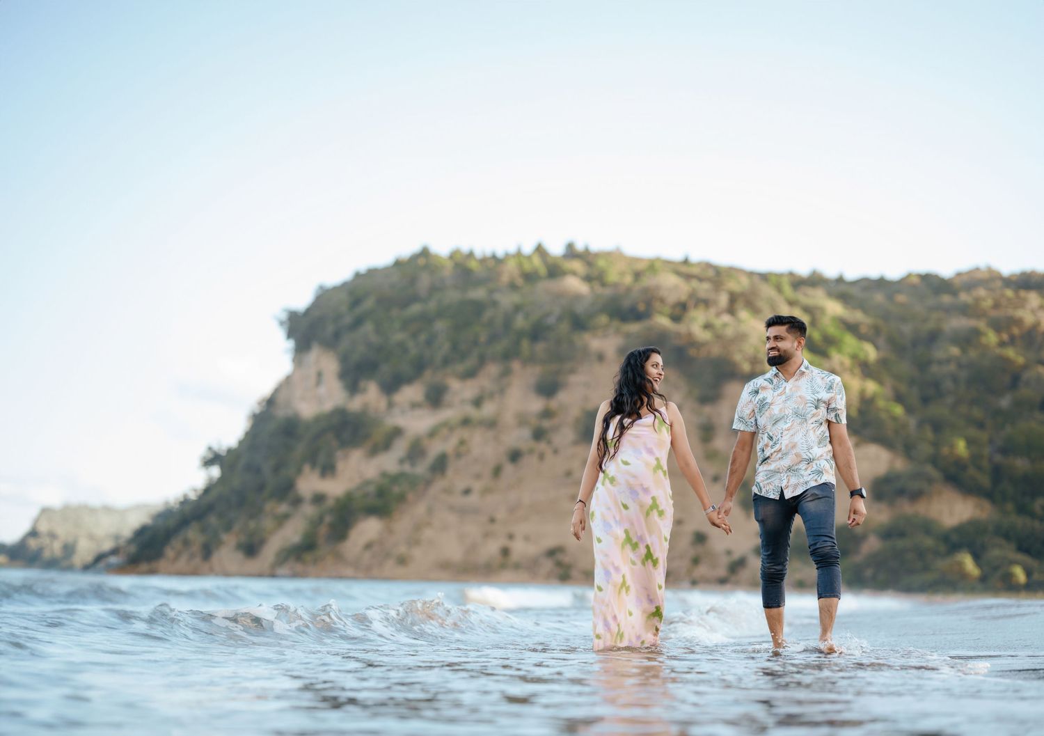 A couple walks along a beach shoreline with cliffs and mountains visible in the background during golden hour.